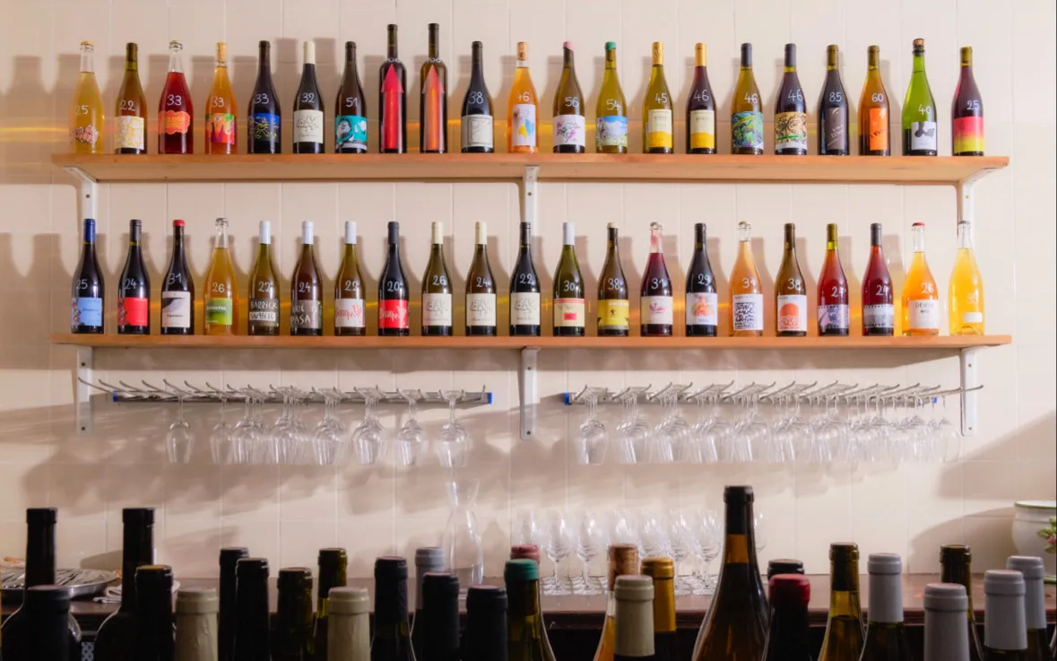 Botellas de vino natural ordenadas en dos estantes, con copas colgadas debajo.