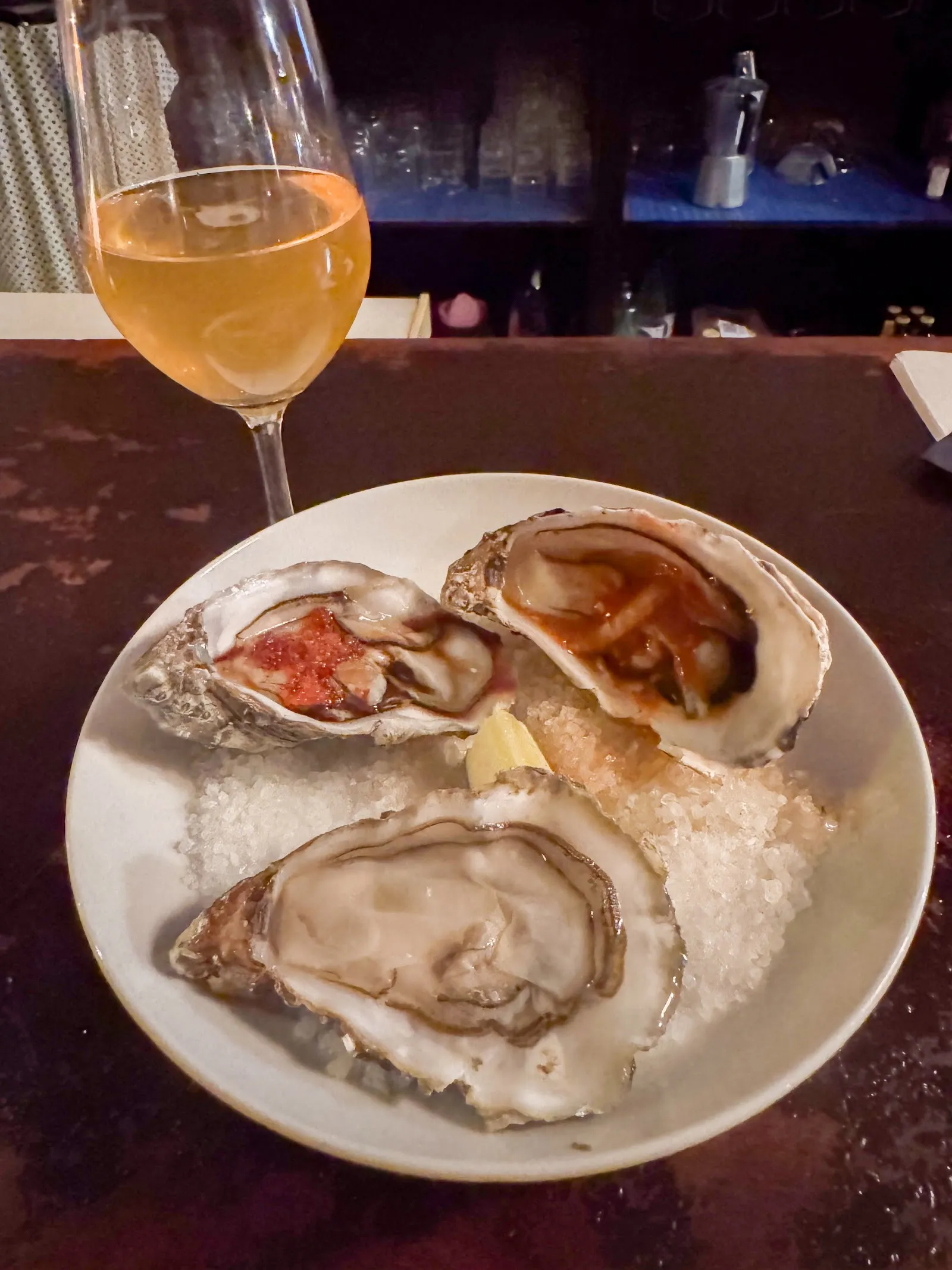 Tres ostras sobre hielo y una copa de vino naranja en la barra.